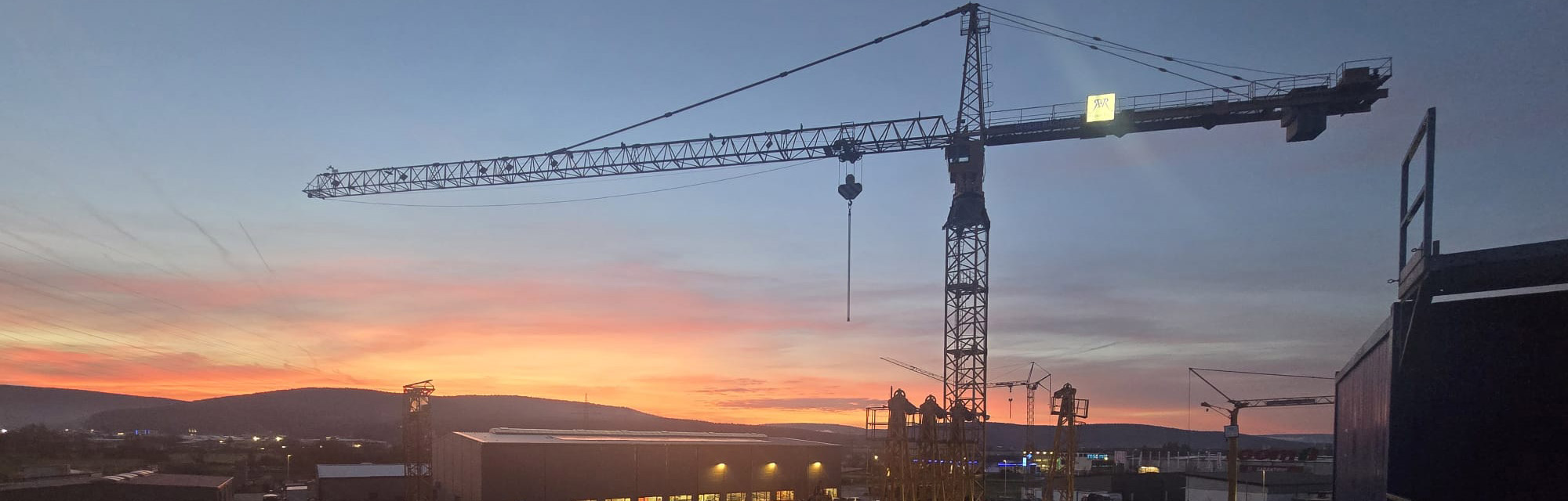 Ein Kran und im Hintergrund geht die Sonne unter. Der Himmel verfärbt sich rosa-orange.