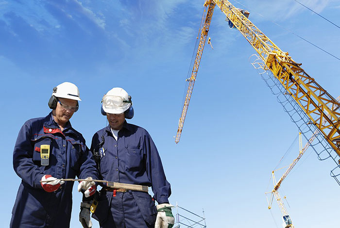 Zwei Bauarbeiter mit einem Zollstock, im Hintergrund ein Kran und blauer Himmel.